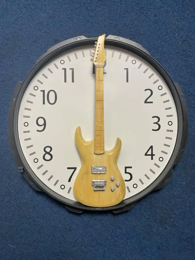 A wall clock on a blue background, its hands replaced by a miniature wooden electric guitar — the neck pointing to twelve and the body hanging below the centre.