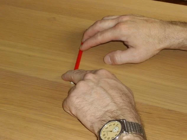 Two hands at a wooden table demonstrating the pencil exercise: one hand holds a pencil flat against the table while the index and middle fingers of the other hand alternate picking strokes against it.